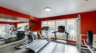 Located at 8900 Chimney Rock Rd in Houston, Texas, this dedicated fitness center delivers a well-equipped workout environment designed to support a full range of exercise routines. The space is defined by its bold red accent walls, which create an energetic and motivating atmosphere throughout. Durable rubber flooring covers the entire area, providing a practical surface suited for both cardio and strength training activities.

The cardio zone features multiple treadmills and an elliptical trainer, positioned along a windowed wall that allows abundant natural light to fill the room and offers views of the outdoor surroundings. Full-length mirrors along one wall visually expand the space and assist users during workouts. Motivational phrases, including "Whatever You Do, Do Not Give Up" and "Don't Count the Days, Make the Days Count," are displayed as wall decals, reinforcing an encouraging environment.

The strength training area is well-stocked with a multi-station cable machine, a lat pulldown, a leg press, a seated row, and additional weight machines, all finished in white with black upholstery. The facility is illuminated by a combination of flush-mount and recessed ceiling lights. French doors on one side provide direct access to the outdoor area, enhancing convenience and ventilation. The overall layout is functional, open, and thoughtfully organized to accommodate a variety of fitness needs.