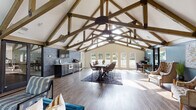 This single-story space is defined by its striking architectural centerpiece: soaring vaulted ceilings supported by a series of exposed wood trusses that span the full length of the interior. The open-plan layout seamlessly integrates several functional zones, creating a versatile and welcoming environment throughout.

The kitchenette is outfitted with slate-grey cabinetry, stainless steel appliances, and a light countertop, set against a dark tile backsplash wall that adds a bold, industrial contrast. Adjacent to the kitchen, a central dining area features a substantial dark wood table surrounded by woven rattan-style chairs, anchored by a patterned area rug on rich wood-look flooring that runs throughout the entire space.

Large French doors along the rear wall flood the interior with natural light and offer views of the surrounding greenery, reinforcing the open, airy atmosphere. A dedicated lounge area near the entrance features a vibrant teal sofa, patterned accent chairs, and a rustic wood-paneled accent wall, providing a relaxed gathering spot.

A quieter seating nook is appointed with tufted wingback chairs set against a deep teal accent wall adorned with abstract artwork and flanked by gold-toned side tables and floor lamps, lending an elegant, curated touch to the overall ambiance.