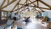 This single-story space is defined by its striking architectural centerpiece: soaring vaulted ceilings supported by a series of exposed wood trusses that span the full length of the interior. The open-plan layout seamlessly integrates several functional zones, creating a versatile and welcoming environment throughout.

The kitchenette is outfitted with slate-grey cabinetry, stainless steel appliances, and a light countertop, set against a dark tile backsplash wall that adds a bold, industrial contrast. Adjacent to the kitchen, a central dining area features a substantial dark wood table surrounded by woven rattan-style chairs, anchored by a patterned area rug on rich wood-look flooring that runs throughout the entire space.

Large French doors along the rear wall flood the interior with natural light and offer views of the surrounding greenery, reinforcing the open, airy atmosphere. A dedicated lounge area near the entrance features a vibrant teal sofa, patterned accent chairs, and a rustic wood-paneled accent wall, providing a relaxed gathering spot.

A quieter seating nook is appointed with tufted wingback chairs set against a deep teal accent wall adorned with abstract artwork and flanked by gold-toned side tables and floor lamps, lending an elegant, curated touch to the overall ambiance.