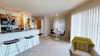 This 1-bedroom, 1-bathroom residence offers a well-organized single-floor layout designed to maximize comfort and functionality. The open-concept living and dining area is anchored by plush carpeting and filled with natural light streaming through large vertical-blind windows and a sliding glass door, creating a bright and welcoming atmosphere. A compact dining nook accommodates a round table with modern chairs, making it an ideal space for casual meals or entertaining. The kitchen features a raised breakfast bar with seating for two, granite-style countertops, and warm wood cabinetry, seamlessly connecting the cooking space to the living area. Track lighting throughout provides ample illumination and adds a contemporary touch to the textured ceilings. The layout ensures a smooth flow between spaces, offering both an open feel and clearly defined areas for living, dining, and cooking. The bedroom provides a private retreat, while the bathroom rounds out this efficient and inviting floor plan. Overall, this residence balances modern design elements with practical living features, making it a comfortable and well-appointed home.