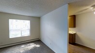 This single-floor space presents a straightforward and functional layout. The main living area features wall-to-wall carpet flooring and is finished with a textured popcorn ceiling. A large window along one wall allows natural light to filter into the room, casting bright patterns across the floor and creating an airy atmosphere. A baseboard heating unit runs beneath the window, providing an efficient heating solution. The walls are painted in a neutral, light tone, lending the space a clean and open feel.

A partial dividing wall separates the living area from the adjacent kitchen, which is visible from the main room. The kitchen features warm-toned wood cabinetry, light countertops, and hard surface flooring, offering a practical contrast to the carpeted living space. The cabinetry provides ample storage, and the layout appears well-suited for everyday use.

Overall, the space maintains a simple, utilitarian character with neutral finishes throughout. The combination of natural light, baseboard heating, and the open connection between the living and kitchen areas contributes to a functional and livable environment.