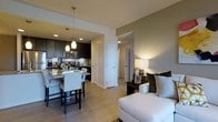 This single-floor residence offers a thoughtfully designed open-concept layout that brings together the living, dining, and kitchen areas in one cohesive, flowing space. The kitchen is outfitted with dark espresso cabinetry, granite countertops, and stainless steel appliances, including a built-in microwave and range. A breakfast bar with seating provides a casual dining option directly adjacent to the kitchen, illuminated by stylish pendant lighting that hangs from the ceiling above. The light hardwood flooring extends throughout the main living area, contributing to a warm and inviting ambiance. The living space is bright and comfortable, featuring neutral-toned walls accented by a warm sage green, complemented by recessed lighting and a classic table lamp that adds a cozy glow. A hallway visible from the main living area leads to additional rooms, offering a sense of depth and privacy within the layout. White interior doors and clean trim work throughout lend a crisp, modern finish. Overall, the space balances contemporary design elements with functional living, making it an ideal environment for both everyday comfort and easy entertaining.
