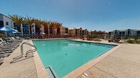 This outdoor amenity space centers around a generously sized swimming pool with clear, blue-tinted water and marked depth indicators along its edges. The pool is framed by a broad, textured concrete sundeck that accommodates a substantial collection of sling-style lounge chairs arranged in organized rows, providing ample space for sunbathing and relaxation. Several blue market umbrellas are positioned throughout the deck, offering shaded seating options during peak sun hours.

The pool area is fully enclosed by a metal security fence, ensuring a safe and private environment. Beyond the fencing, desert-adapted landscaping features gravel ground cover, young trees, native shrubs, and ornamental plantings that contribute to a low-maintenance, arid-climate aesthetic. Tall palm trees punctuate the perimeter, adding a tropical character to the setting.

A clubhouse or amenity building is visible at one end of the pool area, serving as an architectural anchor to the space. The surrounding multi-story residential buildings provide a backdrop, visible across well-maintained green lawns and walking paths. Under clear blue skies, the space benefits from abundant natural sunlight throughout the day, creating an open, resort-inspired atmosphere ideal for outdoor leisure.