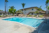 Located at 2230 W Orange Ave in Anaheim, California, this outdoor communal space centers around a generously sized swimming pool that serves as the focal point of the property. The pool area is surrounded by a concrete deck furnished with lounge chairs, tables, and seating, providing ample space for relaxation and recreation under the Southern California sun. A safety ring is visibly mounted near the pool, underscoring the area's well-maintained amenities.

The landscaping throughout the space is lush and thoughtfully arranged, featuring tall, mature palm trees and Norfolk Island pines that rise prominently against the clear blue sky, lending a classic California resort ambiance. Manicured shrubs and flowering plants border the fenced perimeter, adding color and greenery to the environment.

The property's single-story buildings wrap around a portion of the pool area, featuring brown-trimmed facades, large windows, and covered entryways that face the courtyard. A two-story structure with an exterior staircase is also visible at the rear, offering additional depth to the complex. The wrought iron fencing that encloses the pool area provides both security and an open, airy feel. Overall, the space offers a well-appointed, resort-style outdoor retreat ideally suited to the warm Anaheim climate.