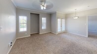 This single-story residence offers a well-organized layout with distinct living, kitchen, and bedroom areas connected by a cohesive neutral color palette. The kitchen is equipped with white cabinetry featuring light wood trim, granite-style countertops, a mosaic tile backsplash, a stainless steel double sink, and black appliances including a range and refrigerator. A breakfast bar peninsula separates the kitchen from the adjacent living area, enhancing the open flow between spaces. The living area features crown molding, a ceiling fan with light fixture, plush wall-to-wall carpeting, and multiple windows alongside a set of French doors that invite natural light and provide outdoor access. The bedrooms continue the carpeted flooring and neutral tones, with ceiling fans and windows overlooking greenery. The primary bathroom includes a wide vanity with a large mirror, built-in lighting, wood-look flooring, and access to a walk-in closet. A secondary bathroom features a tub-shower combination with a two-tone tile surround and open shelving for storage. Throughout the residence, white trim and baseboards provide clean visual definition, contributing to a consistent and functional aesthetic.