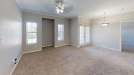 This single-story residence offers a well-organized layout with distinct living, kitchen, and bedroom areas connected by a cohesive neutral color palette. The kitchen is equipped with white cabinetry featuring light wood trim, granite-style countertops, a mosaic tile backsplash, a stainless steel double sink, and black appliances including a range and refrigerator. A breakfast bar peninsula separates the kitchen from the adjacent living area, enhancing the open flow between spaces. The living area features crown molding, a ceiling fan with light fixture, plush wall-to-wall carpeting, and multiple windows alongside a set of French doors that invite natural light and provide outdoor access. The bedrooms continue the carpeted flooring and neutral tones, with ceiling fans and windows overlooking greenery. The primary bathroom includes a wide vanity with a large mirror, built-in lighting, wood-look flooring, and access to a walk-in closet. A secondary bathroom features a tub-shower combination with a two-tone tile surround and open shelving for storage. Throughout the residence, white trim and baseboards provide clean visual definition, contributing to a consistent and functional aesthetic.