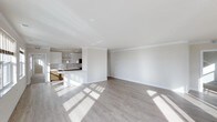 This single-storey space showcases a contemporary open-plan layout that seamlessly connects the living area to the kitchen. The expansive living space is bathed in natural light, which streams through windows and casts warm patterns across the light grey hardwood flooring. Crisp white walls and white crown moulding trim throughout lend a clean, modern aesthetic, while recessed ceiling lighting adds a subtle, functional touch.

The kitchen is thoughtfully designed with white cabinetry, a neutral stone-effect tile backsplash, and dark countertops. A breakfast bar-style peninsula defines the boundary between the kitchen and living area, offering both additional workspace and casual seating potential. Pendant lighting hangs above the peninsula, adding a refined focal point to the kitchen zone. Stainless steel appliances are integrated seamlessly into the cabinetry.

The generous proportions of the living area provide ample flexibility for furniture arrangement. The overall ambiance of the space is light, airy, and modern, making it well-suited for a variety of uses.
