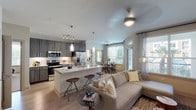 This single-floor residence at 11576 Pearland Pkwy in Houston, Texas offers a thoughtfully designed open-concept layout that seamlessly connects the kitchen, dining, and living areas. The gourmet kitchen is equipped with stainless steel appliances, dark wood cabinetry with sleek hardware, a subway tile backsplash, and a large granite-topped island with bar seating and pendant lighting overhead — ideal for both cooking and entertaining. Track lighting and under-cabinet illumination add to the kitchen's functional appeal. The open living room features light wood-style flooring, a ceiling fan, and multiple windows that invite generous natural light and views of the surrounding greenery. The dining area accommodates a round table beneath a stylish chandelier, framed by large windows with sheer patterned curtains. Both bedrooms are carpeted and well-lit with natural light from oversized windows; the primary bedroom includes its own ceiling fan and ample space. The bathroom is clean and modern, featuring a soaking tub with tiled surround, granite countertops, and wood-toned cabinetry. Neutral tones and contemporary finishes carry throughout, creating a cohesive and welcoming atmosphere across the entire residence.