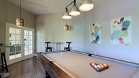 This single-floor space is centered around a well-appointed billiards room designed for leisure and social enjoyment. The room's standout feature is a full-size pool table with a neutral felt surface and a rustic wood base, perfectly positioned beneath a trio of industrial-style pendant lights that cast a warm, focused glow over the playing surface. An additional smaller pendant light adds a decorative accent to the overall lighting arrangement. The walls are finished in a soft, muted gray-green tone, lending the room a calm and inviting atmosphere. Wood-style flooring runs throughout, adding warmth and a contemporary edge to the space. Along one wall, a high-top bar table is flanked by two sleek black bar stools, providing a casual seating area for spectators or those awaiting their turn. A colorful abstract canvas artwork adorns the wall, introducing a vibrant artistic element that contrasts tastefully with the neutral palette. Natural light filters in through a glass-paneled door, connecting the interior to the outdoors and enhancing the overall brightness of the room. The space is thoughtfully designed to encourage social interaction and relaxed recreation.
