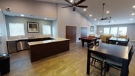 This single-floor space is designed as a multifunctional gathering and recreation area, combining entertainment amenities with comfortable lounge space in an open, flowing layout. The most striking architectural feature is the soaring vaulted ceiling, which lends the space a grand, airy quality enhanced by recessed lighting and large modern ceiling fans that ensure comfort throughout.

The space is anchored by a substantial wet bar with rich dark cabinetry and a light-toned granite countertop, complemented by a built-in sink. A window above the bar area allows natural light to filter in. Adjacent to the bar, a high-top dining table with bar-height seating provides a casual spot for socializing.

A full-size billiards table with a bold red felt surface serves as the centerpiece of the recreation zone, offering an immediate focal point. Beyond it, the space transitions into a relaxed lounge area furnished with upholstered seating and warm ambient lighting, creating a comfortable retreat.

Warm hardwood-style flooring runs throughout, unifying the various zones cohesively. Dark wood double doors provide access to additional rooms, maintaining a consistent design aesthetic across the space.