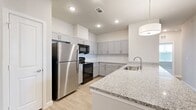 This well-appointed home offers five bedrooms, four bathrooms, and three closets across a thoughtfully designed single-level layout. The open-concept kitchen serves as a central highlight, showcasing granite countertops, a large peninsula with an undermount sink, grey shaker-style cabinetry extending to the ceiling, stainless steel appliances including a full-size refrigerator, and light wood-style tile flooring that flows throughout the main living areas. Recessed lighting and a modern pendant light over the peninsula add a clean, contemporary feel. A hallway with a built-in closet leads seamlessly from the entry into the main living space, enhancing both function and flow. The bedrooms feature plush carpeting, ceiling fans, and large windows with blinds that invite natural light and outdoor views. Multiple rooms benefit from dual-window configurations, further brightening the interiors. The bathrooms are finished with a clean, neutral palette, including a full tub-shower combo with tiled surround and brushed nickel fixtures. Storage is well addressed with multiple walk-in closets equipped with wire shelving systems and carpeted floors, offering ample organization space. The overall aesthetic is modern and neutral, with soft grey and beige tones throughout creating a calm, move-in-ready environment.