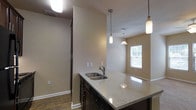 This single-floor residence offers a thoughtfully designed open-concept layout that seamlessly connects the kitchen and dining areas, creating a functional and inviting living environment. The kitchen serves as a central hub, anchored by a spacious island with light-toned granite countertops and a stainless steel sink. Dark cabinetry beneath the island provides ample storage while adding a rich contrast to the lighter surfaces. Three pendant lights hang above, offering both task lighting and a decorative focal point that enhances the kitchen's modern aesthetic. The flooring throughout is a warm hardwood-style material that adds continuity and a sense of warmth to the space. Neutral, beige-toned walls keep the interior feeling bright and versatile, complementing the overall color palette. A window in the dining area allows natural light to filter in, brightening the space and providing a connection to the outdoors. Recessed ventilation is thoughtfully integrated into the ceiling, contributing to a clean and polished finish. The layout is efficiently organized, with a hallway visible from the kitchen that leads to additional areas of the unit, suggesting a well-structured and practical floor plan suited for everyday living.