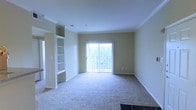 This single-floor residence offers a thoughtfully designed living space that blends comfort and functionality. The main living area is a standout feature, showcasing plush wall-to-wall carpeting that adds warmth underfoot and a welcoming ambiance throughout. Crown molding runs along the ceiling, lending a refined, polished touch to the space.

One wall is anchored by a built-in shelving unit, providing both display and storage possibilities while serving as a distinctive architectural detail. Neutral, warm-toned walls create a versatile backdrop adaptable to a variety of interior styles.

Natural light flows generously into the living area through a large sliding glass door, which opens to a private outdoor balcony enclosed by a decorative wrought-iron railing. The balcony offers a view of lush greenery beyond, bringing a pleasant connection to the outdoors. The sliding door allows for easy indoor-outdoor transition and fills the interior with a bright, airy feel.

The overall design is clean and understated, with a layout that maximizes usable floor space. The combination of natural light, neutral finishes, built-in storage, and outdoor access makes this a practical and comfortable single-floor living space.