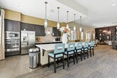 Located at 11405 Central Ave in Chino, CA, this single-story community clubhouse offers a generously sized, open-plan interior designed for gathering and socializing. The space is defined by its warm, contemporary aesthetic, combining teal and neutral wall tones with wood-look flooring, plush carpeting, and carefully curated furnishings throughout.

The main lounge area features multiple seating arrangements, including a large sectional sofa, mid-century modern accent chairs in mustard yellow, and intimate conversation groupings — all positioned beneath recessed lighting and an elegant orb chandelier. Framed artwork and decorative shelving add a refined, welcoming character to the space.

A dedicated dining area accommodates several round tables with upholstered chairs, bathed in natural light streaming through large windows dressed with patterned drapes and plantation shutters. Direct access to an outdoor courtyard is provided through wide glass double doors.

The fully equipped community kitchen is a standout feature, with dark cabinetry, mosaic tile backsplash, stainless steel appliances including a refrigerator and double oven, a long island with a white quartz countertop, and pendant lighting overhead. A row of teal bar stools lines the island, creating an inviting space for casual dining. The overall ambiance is bright, polished, and thoughtfully designed for communal use.