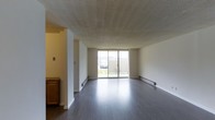 This single-floor space offers a straightforward and functional layout designed for comfortable everyday living. The main living area is open and generously proportioned, featuring dark hardwood-style flooring that runs throughout and provides a sleek, modern contrast to the light-toned walls. A textured ceiling adds subtle character to the space overhead. At the far end of the living area, a large sliding glass door fills the room with natural light and offers a direct view to the outdoors, creating a bright and airy atmosphere. Baseboard heating units line the walls, ensuring year-round comfort. A partial wall divides the living area from an adjacent space, adding a sense of definition without fully closing off the floor plan. To the side, a glimpse of cabinetry with a warm wood finish hints at a kitchen area, suggesting a practical transition between living and cooking spaces. The overall ambiance is clean, neutral, and move-in ready, offering a blank canvas well-suited for a variety of interior styles and furnishing arrangements.