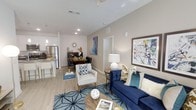 This single-floor residence offers an open-concept layout that seamlessly connects the living, dining, and kitchen areas into one cohesive and functional space. The living area serves as the heart of the home, featuring light-toned luxury vinyl plank flooring, recessed lighting, and a ceiling fan that contribute to a bright and airy atmosphere. The space is anchored by a bold navy blue sofa, a coordinating accent chair, and a geometric area rug, all of which add a vibrant, contemporary character. Colorful wall art enhances the visual appeal and gives the space a curated, modern aesthetic. The dining area sits just beyond the living space and features a dark wood table with seating for several guests, illuminated by a decorative sunburst wall accent. The kitchen is well-appointed with stainless steel appliances, including a full-size refrigerator, light cabinetry, and a stone-finish countertop with bar seating — ideal for casual dining or entertaining. Neutral warm-toned walls run throughout, tying the spaces together cohesively. The overall ambiance is polished and inviting, blending modern design elements with comfortable, livable touches that make this a highly functional and visually appealing space.