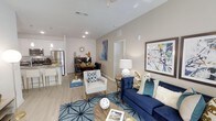 This single-floor residence offers an open-concept layout that seamlessly connects the living, dining, and kitchen areas into one cohesive and functional space. The living area serves as the heart of the home, featuring light-toned luxury vinyl plank flooring, recessed lighting, and a ceiling fan that contribute to a bright and airy atmosphere. The space is anchored by a bold navy blue sofa, a coordinating accent chair, and a geometric area rug, all of which add a vibrant, contemporary character. Colorful wall art enhances the visual appeal and gives the space a curated, modern aesthetic. The dining area sits just beyond the living space and features a dark wood table with seating for several guests, illuminated by a decorative sunburst wall accent. The kitchen is well-appointed with stainless steel appliances, including a full-size refrigerator, light cabinetry, and a stone-finish countertop with bar seating — ideal for casual dining or entertaining. Neutral warm-toned walls run throughout, tying the spaces together cohesively. The overall ambiance is polished and inviting, blending modern design elements with comfortable, livable touches that make this a highly functional and visually appealing space.