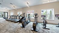 Located at 2031 Maywill St in Richmond, VA, this well-appointed fitness center offers a comprehensive, single-floor workout facility designed with both function and aesthetics in mind. The space is anchored by a distinctive geometric-patterned carpet that runs throughout, lending a contemporary feel to the environment. Warm-toned structural columns punctuate the open floor plan, complementing the clean white walls and modern linear lighting fixtures overhead.

The cardio zone features a strong lineup of equipment, including multiple treadmills, stair climbers, ellipticals, and stationary cycling bikes, many equipped with individual digital display screens. Large windows line one wall, flooding the space with natural light and offering views of the surrounding exterior, while ceiling fans contribute to a comfortable workout atmosphere.

The strength and functional training area is equally well-equipped, with a full dumbbell rack, cable machines, a multi-station strength system, and additional resistance equipment. Wall-mounted flat-screen televisions, framed in warm wood surrounds, are positioned for easy viewing from the training area. A graphic accent wall and built-in cabinetry add refined detail to this section of the facility. The overall design balances practicality with a polished, modern aesthetic, creating an inviting environment for a full range of fitness activities.