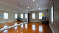 This single-floor fitness studio is thoughtfully designed to support a wide range of movement-based activities, from yoga and Pilates to dance and personal training. The room's most striking feature is the floor-to-ceiling mirror spanning an entire wall, creating a sense of expanded space while allowing for form and technique monitoring during workouts. The rich, polished hardwood floors provide both aesthetic warmth and a durable, low-impact surface suited for exercise. Recessed lighting is evenly distributed across the smooth ceiling, accented by classic crown molding that adds an elegant touch to the otherwise functional space. Multiple windows fitted with white plantation shutters line the walls, inviting generous natural light and offering a bright, energizing atmosphere. The neutral, light-toned walls contribute to an open and airy feel. A medicine ball rack stocked with various weighted balls is positioned along one wall, while exercise balls in multiple sizes add to the room's well-equipped nature. The overall ambiance is clean, spacious, and purposeful, combining the refined details of a well-appointed interior with the practical demands of a dedicated home fitness environment.