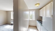 This 1-bedroom, 1-bathroom unit is designed with a straightforward, efficient layout across a single floor. The kitchen is a compact, galley-style space featuring white cabinetry extending to the ceiling for ample storage, a gas range, and a refrigerator. Light-toned tile flooring and a window framing views of surrounding trees keep the kitchen feeling bright and practical.

The living area and bedroom are finished with wall-to-wall carpet in a neutral gray tone, providing a comfortable, cohesive feel throughout. The bedroom features a large window that fills the room with natural light and offers pleasant views of mature trees, along with a radiator unit for heating. The living area similarly benefits from natural light and provides a clean, open canvas for furnishing.

The bathroom includes a full bathtub with shower, white surround walls, and a window that introduces natural light and ventilation. Radiator heating is present in the main living spaces, and neutral beige-toned walls are consistent throughout the unit. The overall ambiance is simple and functional, with greenery visible from multiple windows providing a calm, natural backdrop.
