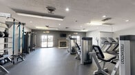 This fitness center offers a generously sized, single-floor workout space designed to accommodate a full range of exercise activities. The facility is outfitted with durable rubber flooring throughout, providing a safe and functional surface for all types of training. A comprehensive selection of equipment is distributed across the open layout, including multiple treadmills, elliptical machines, and stair climbers for cardiovascular workouts, as well as a variety of strength training machines and a functional rig system complete with cable attachments, resistance bands, and medicine balls. A dedicated shelving unit organizes free weights and accessories for easy access. The space is finished with neutral grey walls and a textured ceiling fitted with a combination of recessed lighting and fluorescent fixtures, ensuring bright, even illumination throughout. A standout architectural feature is the traditional-style fireplace along the main wall, flanked by large glass doors that invite natural light and provide views to an outdoor area. A wall-mounted television is positioned above the fireplace, adding an entertainment element to the workout environment. The overall ambiance is clean, modern, and purposefully designed to support an active and health-focused lifestyle.