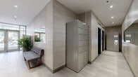 Located at 1420 Rue Crescent in Montréal, this lobby hallway serves as the primary entry and circulation space for the building. The interior design reflects a cohesive contemporary aesthetic, with light-toned horizontal wall cladding complemented by contrasting dark tile accents along key feature walls. Large-format porcelain floor tiles in a neutral stone-like finish run throughout, contributing to a clean and polished appearance.

A prominent bank of individual mailboxes lines one wall, finished in brushed metal and illuminated by recessed pot lighting above. Adjacent to these, tall parcel locker units provide secure package storage for residents. Elevator access is integrated seamlessly into the hallway, framed by the same refined wall paneling used throughout the space.

Near the glazed entrance doors, natural light filters into the lobby, offering views onto Rue Crescent. A tufted upholstered bench provides a comfortable waiting area, accompanied by potted tropical plants that introduce a natural element to the otherwise sleek environment. A wall-mounted mirror further enhances the sense of openness. The staircase, finished with dark tile risers, connects to additional building levels. Recessed ceiling lighting ensures the entire hallway is well-lit, creating a welcoming and functional environment.