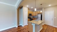 This single-floor apartment at Olde Towne Apartments offers a thoughtfully designed living space with a blend of modern finishes and functional layout. The kitchen serves as a central highlight, featuring warm honey-toned wood cabinetry, granite countertops, and a full suite of stainless steel appliances including a refrigerator, electric range, and built-in microwave. A peninsula island with a double-basin sink and pendant lighting overhead adds both workspace and a casual dining option, making the kitchen ideal for everyday cooking and entertaining. Rich hardwood flooring flows throughout the space, lending warmth and continuity to the open layout. Arched architectural detailing frames the transition between the kitchen and the adjoining living area, adding a touch of classic character to the otherwise contemporary design. Neutral warm-toned walls complement the natural wood finishes and create a welcoming ambiance. Recessed lighting illuminates the space efficiently, while pendant lights above the island provide focused task lighting. A closet off the kitchen area provides convenient storage access. Overall, the apartment combines practical amenities with tasteful design elements for comfortable everyday living.