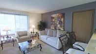 This single-floor space offers a thoughtfully designed interior that blends comfort and contemporary style. The living area serves as the centerpiece of the home, featuring plush seating arranged around a glass-topped coffee table with ornate metalwork detailing. A bold accent wall painted in a deep gray tone provides a striking contrast to the surrounding neutral palette, and is adorned with a vibrant piece of abstract artwork that draws the eye and adds a splash of color to the room. Soft, light-colored carpeting underfoot contributes to the warm and inviting ambiance throughout the space. Natural light filters in through vertical blinds on a large window, brightening the room and creating an airy feel. Lush indoor greenery adds a refreshing, organic touch to the décor. Adjacent to the living area, a dining space with dark-framed chairs is seamlessly integrated, making the layout well-suited for both relaxing and entertaining. The overall design is characterized by a harmonious mix of soft neutrals and carefully chosen accent pieces, resulting in a space that feels both polished and welcoming.