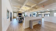 This expansive single-story clubhouse offers a thoughtfully designed community gathering space spread across a generous floor plan. The architectural character is defined throughout by distinctive coffered ceilings with crisp white beams, complemented by warm hardwood-style flooring that unifies the entire space.

The main lounge area serves as the heart of the clubhouse, featuring a stone fireplace, wall-mounted television, and comfortable seating arrangements beneath elegant chandelier lighting. An adjacent open area provides additional flexible seating, bathed in natural light from large windows fitted with layered roller shades.

The fully equipped kitchen is outfitted with white shaker cabinetry, granite countertops, stainless steel appliances including a french-door refrigerator and built-in microwaves, and a central island with bar seating—ideal for community events and gatherings.

A dedicated business center offers built-in desk workstations, modern seating, and curated artwork, providing a focused environment for residents. A separate meeting room features a marble-top round table and framed art displays. Additional spaces include a game room with a foosball table and a formal dining area with a large farmhouse table and wall-mounted television, completing this well-appointed clubhouse.