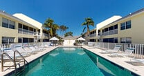 Located in the heart of Cape Coral, Florida, this inviting outdoor pool area offers a classic Florida resort-style experience. The centerpiece is a generously sized swimming pool with crystal-clear turquoise water, accessible via traditional handrail-equipped entry steps, making it welcoming for all users. The pool is enclosed by a white metal safety fence, ensuring a secure and well-defined recreational space.

Surrounding the pool is an expansive concrete sundeck furnished with multiple lounge chairs and seating arrangements, providing ample space for relaxation under the abundant Southwest Florida sunshine. Shaded areas are created by patio umbrellas positioned around the deck, offering a cool retreat from the sun. Outdoor tables and chairs are available for casual gatherings.

A utility or amenity structure is situated at one end of the pool area, featuring restroom or storage facilities, adding convenience for guests. The complex is framed by a two-story residential building with a warm yellow exterior, screened balconies, and a pitched roof, contributing to the community's cohesive architectural style. Mature palm trees and tropical landscaping border the space, enhancing the lush, resort-like ambiance. Under clear blue skies, this pool area embodies the relaxed, sun-drenched lifestyle characteristic of Cape Coral living.