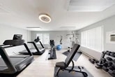 Located at 8684 Drumlin Heights Dr in Baldwinsville, NY, this fitness center offers a fully outfitted workout facility across a single floor. The space is designed with functionality and comfort in mind, featuring light wood-look flooring, soft gray walls, and a vibrant teal accent wall that adds a pop of energy to the environment. Multiple windows allow natural light to fill the room, creating a bright and inviting atmosphere for exercise.

The main workout area is well-stocked with a variety of cardio equipment, including multiple treadmills, a stationary upright bike, a recumbent bike, and an elliptical machine. A multi-station cable and strength machine anchors one side of the room, while a full rack of dumbbells and an adjustable weight bench provide ample free weight options. An exercise ball and wall-mounted mirror round out the functional amenities. A wall-mounted television and sanitization station add to the convenience of the space.

Overhead, a modern circular ceiling fan with integrated lighting provides ventilation and a contemporary aesthetic touch, complemented by flush-mount panel lighting throughout. A dedicated storage closet keeps the area organized. The fitness center is accessible via a paneled white door and is well-suited for a broad range of individual workout routines.