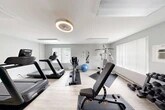 Located at 8684 Drumlin Heights Dr in Baldwinsville, NY, this fitness center offers a fully outfitted workout facility across a single floor. The space is designed with functionality and comfort in mind, featuring light wood-look flooring, soft gray walls, and a vibrant teal accent wall that adds a pop of energy to the environment. Multiple windows allow natural light to fill the room, creating a bright and inviting atmosphere for exercise.

The main workout area is well-stocked with a variety of cardio equipment, including multiple treadmills, a stationary upright bike, a recumbent bike, and an elliptical machine. A multi-station cable and strength machine anchors one side of the room, while a full rack of dumbbells and an adjustable weight bench provide ample free weight options. An exercise ball and wall-mounted mirror round out the functional amenities. A wall-mounted television and sanitization station add to the convenience of the space.

Overhead, a modern circular ceiling fan with integrated lighting provides ventilation and a contemporary aesthetic touch, complemented by flush-mount panel lighting throughout. A dedicated storage closet keeps the area organized. The fitness center is accessible via a paneled white door and is well-suited for a broad range of individual workout routines.