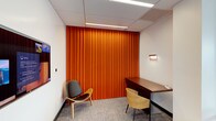 This single-floor meeting room is a thoughtfully designed space that balances function with a distinct visual identity. The most striking feature is the floor-to-ceiling pleated orange fabric wall panel, which serves as a bold focal point and adds warmth and acoustic value to the room. The neutral white walls and drop ceiling with recessed linear lighting create a clean, professional backdrop that allows the vibrant accent wall to stand out. A built-in wooden desk runs along one wall, accompanied by a modern sculpted chair with a light wood frame, providing a practical workstation for participants. A second stylish lounge-style chair with a black seat and bentwood legs adds a touch of contemporary design and flexible seating. A wall-mounted sconce provides supplemental ambient lighting, while a display screen on the adjacent wall supports presentations and collaborative work. The carpeted floor features a subtle multi-toned pattern that complements the overall color palette. The room is well-suited for small meetings, one-on-one consultations, or focused work sessions, combining a professional atmosphere with warm, design-forward aesthetics.
