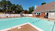 This single-floor community amenity facility is designed to support an active lifestyle, combining a fully outfitted fitness center with a spacious outdoor pool area. The fitness center features durable rubber flooring and textured ceilings throughout, with cool grey walls accented by bold green diagonal stripes that give the space a dynamic, energetic feel. Large windows along one wall allow natural light to flood the room and offer views of the pool area outside. The gym is equipped with a variety of cardio and strength-training machines, including a Spirit treadmill, elliptical trainer, stationary bike, cable weight machine, and a multi-functional Smart Recovery rack stocked with medicine balls and stability balls in multiple sizes. Full-length mirrors on one wall enhance the sense of space. French glass doors at the rear of the fitness center open directly to the outdoors, providing easy access and ventilation. The outdoor pool area features a generously sized, clearly marked swimming pool with lane dividers, surrounded by a textured pool deck. Lounge chairs and umbrella-shaded tables are arranged around the perimeter for relaxation, and a covered pavilion with additional seating sits within the fenced grounds, all set against a backdrop of mature trees.