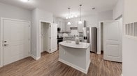 This single-floor residence is thoughtfully designed with a clean, neutral palette and modern finishes throughout. The kitchen serves as a central highlight, showcasing gray shaker-style cabinetry, granite-look countertops, and a full suite of stainless steel appliances including a gas range, microwave, dishwasher, and a French door refrigerator with an ice and water dispenser. A freestanding island with a stone countertop provides additional prep space and casual seating, while pendant lighting and wood-look flooring complete the contemporary feel. The open layout connects the kitchen to the main living area, which continues the warm wood-look flooring and is fitted with a ceiling fan for added comfort. The bedrooms are carpeted and finished in soft gray tones, each equipped with a ceiling fan and windows that allow natural light to filter in, creating a calm and restful atmosphere. The full bathroom features a tub-shower combination, a vanity with granite-look countertops, wood-look flooring, and recessed lighting for a polished finish. Completing the unit's practical amenities is a dedicated laundry closet housing a full-size top-load washer and dryer, along with overhead wire shelving for storage.