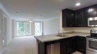 This single-floor residence offers a thoughtfully designed open-concept layout that effortlessly connects the kitchen and living areas. The kitchen is a standout feature, showcasing rich dark cabinetry that contrasts elegantly with light granite countertops. A peninsula with a stainless steel sink and built-in dishwasher provides both additional workspace and a natural boundary between the kitchen and the adjacent living area, making it ideal for entertaining. Crown molding runs along the ceiling throughout, adding a refined architectural detail that elevates the overall aesthetic. The living area is bathed in natural light, courtesy of a large bank of windows and a glass-paned door that opens directly onto a private balcony. Lush tree canopy views visible through the windows create a tranquil, nature-connected ambiance. Soft, neutral carpeting in the living space adds warmth and comfort underfoot, while the clean white walls and light ceiling maintain an airy, open feel. A pendant light fixture anchors the living area, providing both functional illumination and a subtle design accent. The seamless flow between spaces and the abundance of natural light make this residence feel welcoming and spacious throughout.