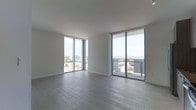This single-floor unit is defined by its striking floor-to-ceiling glass windows that span two exterior walls, flooding the open living space with abundant natural light and framing panoramic views of the city skyline and a shimmering body of water beyond. The corner-facing window configuration maximizes the sense of openness, creating an airy, expansive atmosphere throughout the main living area. Smooth white ceilings complement the clean, minimalist aesthetic, while light gray wood-style flooring runs continuously across the space, adding warmth and a contemporary finish. A sliding glass door provides direct access to a private balcony, extending the living space outdoors and offering an elevated vantage point above the surrounding cityscape. The overall design emphasizes simplicity and openness, with neutral tones and unobstructed sightlines that highlight the dramatic outdoor views as the central feature of the space. The unit's elevated position enhances both the sense of scale and the quality of natural light, making it a visually compelling and well-lit environment throughout the day.