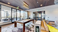 This single-floor amenity space is thoughtfully divided into two distinct yet interconnected areas, together offering a generously sized entertainment and social environment. The main lounge area is defined by light wood-look flooring, bold abstract-patterned area rugs, and a coffered ceiling clad in dark wood paneling—creating a sophisticated contrast against the bright white walls. Floor-to-ceiling glass walls and expansive windows flood the space with natural light and provide views of an adjacent outdoor terrace. A centrally placed billiards table anchors the lounge beneath a striking cascading gold-and-glass pendant chandelier. Surrounding seating includes leather sofas, upholstered lounge chairs, high-top tables, and intimate conversation groupings, catering to a variety of social settings. A large wall-mounted screen enhances the media-viewing experience. The second area functions as a dedicated golf simulator room, featuring a full-swing projection screen, green turf mat, and a ceiling-mounted projector. This room is distinguished by vibrant tropical-print wallpaper, wood-paneled ceilings, and a live-edge wood bar counter with green velvet booth seating, creating a lively, resort-inspired atmosphere. Steel-framed glass partition doors connect both spaces fluidly.