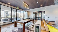 This single-floor amenity space is thoughtfully divided into two distinct yet interconnected areas, together offering a generously sized entertainment and social environment. The main lounge area is defined by light wood-look flooring, bold abstract-patterned area rugs, and a coffered ceiling clad in dark wood paneling—creating a sophisticated contrast against the bright white walls. Floor-to-ceiling glass walls and expansive windows flood the space with natural light and provide views of an adjacent outdoor terrace. A centrally placed billiards table anchors the lounge beneath a striking cascading gold-and-glass pendant chandelier. Surrounding seating includes leather sofas, upholstered lounge chairs, high-top tables, and intimate conversation groupings, catering to a variety of social settings. A large wall-mounted screen enhances the media-viewing experience. The second area functions as a dedicated golf simulator room, featuring a full-swing projection screen, green turf mat, and a ceiling-mounted projector. This room is distinguished by vibrant tropical-print wallpaper, wood-paneled ceilings, and a live-edge wood bar counter with green velvet booth seating, creating a lively, resort-inspired atmosphere. Steel-framed glass partition doors connect both spaces fluidly.