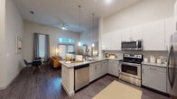 This thoughtfully designed single-floor residence showcases a contemporary open-concept layout that seamlessly connects the kitchen, dining, and living areas. The kitchen is a standout feature, boasting granite countertops, a large peninsula with an undermount sink, sleek white upper cabinetry paired with grey lower cabinets, subway tile backsplash, and a full suite of stainless steel appliances including a refrigerator, range, microwave, and dishwasher. Stylish pendant lighting illuminates the peninsula, adding both function and visual appeal.

The living room impresses with soaring ceilings and oversized windows fitted with blinds and transom windows above, drawing in generous natural light. Dark hardwood-style flooring flows throughout the main living areas, grounding the airy, modern aesthetic. A ceiling fan adds comfort to the open living space.

The bedrooms offer a calm retreat, featuring plush carpeting, tall ceilings, and well-sized windows. The primary bathroom is well-appointed with a granite double vanity, backlit mirror, full soaking tub with tile surround, and dark wood-style flooring, with direct access to a spacious walk-in closet complete with built-in shelving. The overall ambiance throughout the residence is clean, contemporary, and inviting.