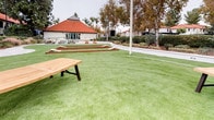 Located at 1699 E Washington St in Colton, California, this well-appointed outdoor recreational space offers a thoughtfully designed environment for leisure and socializing. The centerpiece of the area is a generous expanse of lush artificial turf, providing a clean, low-maintenance green surface that remains vibrant year-round. Multiple bocce ball courts with warm wood-toned frames are laid out across the lawn, inviting friendly competition and outdoor activity.

Wooden picnic tables with attached benches are positioned throughout the turf area, offering comfortable spots for gathering and dining al fresco. A charming gazebo with a distinctive tiered roof serves as a shaded retreat, complemented by seating and a paved surround — ideal for relaxing or hosting smaller gatherings. Raised planter walls with a crisp white finish define the perimeter of the turf area, adding structure to the landscape.

Mature trees, ornamental grasses, and well-maintained shrubs frame the space, providing natural shade and a sense of enclosure. Pathway lighting and a separate covered building with red tile roofing and large glass doors are also visible, suggesting additional amenity or community spaces nearby. The overall ambiance is open, social, and inviting, designed to encourage outdoor engagement in a well-kept community setting.