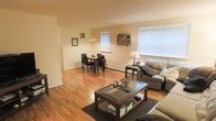 This single-floor space features an open-concept living and dining area that combines functionality with a comfortable, relaxed atmosphere. The warm hardwood floors run throughout, adding a sense of continuity and warmth to the interior. Natural light filters in through multiple windows fitted with horizontal blinds, brightening the neutral, cream-colored walls and contributing to an airy, welcoming feel.

The living area accommodates a cushioned sofa and loveseat arranged around a glass-top coffee table with a lower storage shelf, providing both style and practicality. A side table with a lamp adds soft ambient lighting to complement the natural light. The dining area, situated toward the back of the open space, features a compact table with seating for four, illuminated by a pendant light overhead. A doorway visible on the far left suggests additional rooms beyond the main living space.

Overall, the layout is straightforward and efficient, making good use of the available space. The combination of hardwood flooring, neutral walls, and generous window placement creates a bright, livable environment well-suited for everyday use.