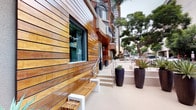 The Preston is a thoughtfully designed single-story commercial space that blends modern aesthetics with warm, natural materials. The entrance is defined by a set of full-glass double doors branded with "The Preston" signage, offering a clear view into the bright, open interior. A secondary entry point features sheer white curtains that soften the transition between the outdoors and interior spaces, adding an airy, refined touch to the façade.

The exterior is a standout architectural feature, clad in horizontal warm-toned hardwood planks that lend a contemporary yet organic character to the building. Large windows framed in sleek dark trim allow natural light to filter in while connecting the interior to the surrounding environment. The outdoor walkway is well-appointed with modern slatted wood benches set on white steel bases, providing functional yet stylish seating.

Tall black cylindrical planters filled with striking succulents and architectural plants are strategically placed along the exterior, adding lush greenery to the hardscaped surroundings. The overall ambiance is clean, polished, and inviting, with a design language that balances natural warmth with contemporary sophistication, making it an appealing destination within its urban setting.