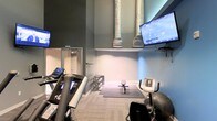 This fitness center offers a thoughtfully designed workout environment across a single level. The space features impressively high ceilings with exposed ductwork rendered in a light finish, lending an industrial character that contrasts with the smooth, neutral-toned walls painted in shades of gray and soft blue. The elevated ceiling height creates an open, airy atmosphere despite the room's intimate footprint.

The facility is equipped with essential cardio machines, including treadmills and a stationary bike, positioned for easy access. A designated stretching and floor exercise area is defined by dark gym mats, accompanied by a dumbbell rack offering a range of free weights for strength training. Multiple wall-mounted flat-screen televisions are strategically placed throughout the space, ensuring visibility from various workout stations for entertainment during exercise sessions.

The flooring consists of light-toned, low-pile carpet suited for fitness use, and recessed lighting provides consistent illumination throughout. A mirrored section near the entrance enhances the sense of space and supports form-checking during workouts. Overall, the gym delivers a clean, functional environment with a modern industrial design.