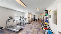 Located at Parkway Village in Grove City, Ohio, this fitness center offers a comprehensive workout facility within a single, open floor. The space features a distinctively patterned carpet in neutral tones with purple accents, bright white walls, and a textured ceiling fitted with recessed fluorescent lighting that keeps the area well-lit throughout.

The cardio zone is equipped with multiple treadmills and elliptical trainers, as well as a stationary bike, all positioned near windows fitted with horizontal blinds that allow natural light to filter in. A multi-station cable machine serves as the centerpiece for strength training, complemented by a dedicated free weight area featuring a tiered dumbbell rack and a full medicine ball rack with a variety of weighted options. Rowing machines are also available, set on protective floor mats.

A wall-mounted flat-screen television adds an entertainment element for those working out, while a large mirror along one wall enhances the sense of space and allows users to monitor their form. The layout is efficiently organized to accommodate a variety of training styles, from cardio and functional fitness to strength and resistance training, making this facility a well-rounded amenity for residents of the Parkway Village community.