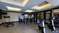 This fitness center offers a thoughtfully designed workout environment that combines functionality with refined aesthetics. The space is anchored by a striking coffered ceiling with recessed lighting panels that provide bright, even illumination throughout, creating an energizing atmosphere for exercise.

The main workout area is outfitted with a selection of modern cardio equipment, including elliptical trainers, a recumbent bike, and additional aerobic machines arranged to allow comfortable movement between stations. A wall-mounted flat-screen television is positioned for easy viewing during workouts.

The flooring consists of durable speckled composite material, ideal for a high-traffic fitness environment. Full-length mirrors along one wall enhance the sense of space and allow for form-checking during exercise. A water cooler station is conveniently accessible near the entrance, ensuring hydration is always within reach.

Large glass-paned doors with dark frames lead to an adjoining area, adding visual depth and connectivity to the overall space. The walls are painted in a cool, neutral tone that complements the dark trim accents and contributes to a clean, professional feel. Overall, this fitness center delivers a well-lit, well-appointed setting suited for a comprehensive cardio workout routine.