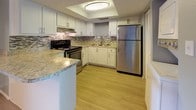 This well-appointed single-floor, 3-bedroom, 2-bathroom home offers a thoughtful layout with a blend of modern updates and comfortable living spaces. The kitchen serves as a functional centerpiece, featuring white shaker-style cabinetry, granite countertops, a mosaic tile backsplash, stainless steel appliances including a refrigerator and electric range, and a recessed ceiling with flush lighting. Light wood-style flooring flows through the main living areas, complementing the soft gray wall tones and textured ceilings throughout. The spacious living room receives natural light through a large window with exterior views and offers ample room for a variety of furniture arrangements. The three bedrooms are each finished with plush carpeting, neutral gray walls, and textured ceilings, with several rooms featuring windows fitted with blinds for natural light and privacy. Both bathrooms are finished consistently with granite-topped white vanities, wood-style tile flooring, and full tub/shower combinations. The overall design maintains a clean, neutral palette, creating a cohesive and move-in ready environment suitable for a range of occupants.