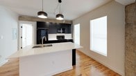 This thoughtfully designed one-bedroom, one-bathroom unit seamlessly merges industrial character with modern aesthetics across a single level. Warm-toned hardwood flooring flows throughout the living spaces, complementing the clean white walls and exposed concrete columns and beams that lend an authentic urban loft feel.

The kitchen is a standout feature, equipped with bold matte black cabinetry, white quartz countertops, and a full suite of black appliances including a refrigerator, range, and microwave. A spacious kitchen island with an undermount sink and brushed gold faucet provides both prep space and casual seating, illuminated by a trio of modern pendant lights. Track lighting and large windows ensure the space remains bright and functional.

The bedroom is generously proportioned, offering multiple windows that flood the room with natural light, and is outfitted with a ceiling fan for added comfort. The bathroom features a dark vanity with a white quartz countertop, a round gold-framed mirror, black hardware accents, and a full tub-shower combination with brushed gold fixtures. An adjoining walk-in closet with built-in shelving adds practical storage.

Additional highlights include a dedicated in-unit laundry closet with a stacked washer and dryer, a private entry door, and an Ameristar HVAC system for efficient climate control.
