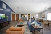 This single-floor community space offers an open, welcoming layout designed for gathering and relaxation. The main lounge area is anchored by vaulted ceilings with a dramatic navy blue accent wall, recessed lighting, and ceiling fans that contribute to an airy, comfortable atmosphere. Durable hardwood-style flooring extends throughout, tying the space together seamlessly.

The lounge is generously sized and thoughtfully furnished with plush sofas, leather accent chairs, and a central coffee table, creating multiple conversation areas suited for both casual socializing and structured gatherings. A wall-mounted television adds an entertainment focal point to the primary seating zone.

An adjacent dining area provides additional seating with tables and chairs, ideal for communal meals or organized activities. Large windows fitted with wood-trimmed blinds line the walls, inviting abundant natural light and offering pleasant outdoor views.

A tiled bar counter with industrial-style bar stools serves as a stylish transition to the kitchen area, which is equipped with wood cabinetry, full-sized appliances, and tile flooring. French doors with warm wood framing provide both interior connectivity and natural light. The overall design blends contemporary and transitional elements, resulting in a functional, inviting space well-suited for community use.