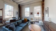 Located at 625 Broadway in the heart of downtown San Diego, this distinctive loft unit (Plan F, #207) masterfully merges historic industrial character with contemporary design. The open-concept living area immediately captivates with its exposed white-painted beam ceilings, original concrete and brick columns, and wide-plank hardwood flooring that runs throughout. Track lighting overhead complements the abundant natural light streaming in through large, classic sash windows that frame views of the surrounding cityscape.

The living space offers a generous layout accommodating both a lounge and dining area, giving the space an airy, loft-like quality. Exposed brick columns punctuate the room, adding warmth and architectural texture that speaks to the building's storied past.

The kitchen is sleek and fully equipped, featuring floor-to-ceiling dark wood cabinetry, light countertops, stainless steel appliances including a refrigerator, range, dishwasher, and microwave, all set beneath soaring ceilings that reinforce the loft aesthetic.

The bedroom is intimate and thoughtfully appointed, with a prominent exposed brick column beside a bright window creating a striking focal point. The bathroom is clean and modern, with dark wood vanity cabinetry, a white countertop, large mirror, and an adjacent closet, rounding out this well-designed urban retreat.